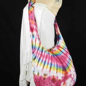May include: A tie-dye cotton shoulder bag with a circular pattern in shades of pink, white, blue, yellow, and green. The bag has a wooden button closure.