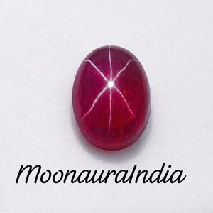 Puede incluir: Una piedra preciosa de rubí estrella de color rojo intenso y forma ovalada que presenta un patrón de estrella de seis rayos en su superficie lisa y pulida. La gema se muestra sobre un inserto de espuma blanca en un estuche cuadrado, con el texto MoonauraIndia impreso debajo.