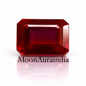 Può includere: Una gemma di rubino sfaccettata e rettangolare, di colore rosso intenso. La gemma è posta su uno sfondo bianco, con il testo "MoonAuraindia" in basso. Le sfaccettature della gemma riflettono la luce, creando un effetto brillante.
