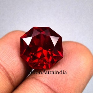 Pode incluir: Uma pedra preciosa facetada, octogonal, vermelho escuro. A gema é segurada em uma mão, com as facetas refletindo a luz. A cor da gema é um vermelho rico e saturado, e o corte cria múltiplos pontos de reflexão.