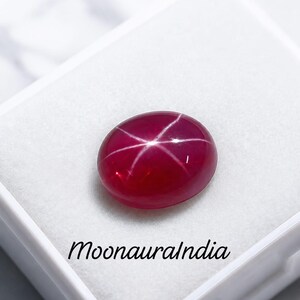 May include: A deep red oval star ruby gemstone featuring a distinct six-rayed asterism effect across its smooth cabochon surface. The gemstone is displayed inside a white box with the text MoonauraIndia printed in black script on the base.
