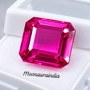 May include: A vibrant, square-cut hot pink gemstone with faceted surfaces that reflect light, displayed inside a white plastic jewellery box. The text Moonauraindia is printed in black on the white fabric insert beneath the gemstone.