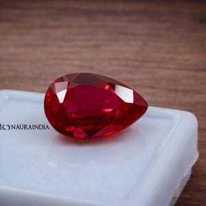 May include: A faceted, pear-shaped, deep red gemstone rests on a white surface. The gem is highly polished, reflecting light and showcasing its brilliant cut. The background is a wooden surface. The text "MONAURAINDIA" is visible on the white surface.