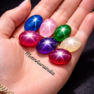 May include: A hand displaying seven oval star gemstones in shades of blue, pink, green, purple, and red. Each gem features a star-shaped light effect. The text "MoonAuraindia" is visible on the hand.