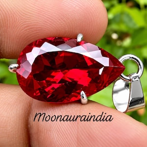 Puede incluir: Un colgante de piedra preciosa roja tallada en forma de pera, montado en un engaste de metal plateado con una anilla a juego. El texto Moonauraindia está impreso en letra cursiva negra debajo de la piedra. El colgante se sostiene en la mano.