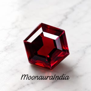 May include: A deep red, hexagonal-cut gemstone with sharp, faceted edges that reflect light. The stone displays a rich, saturated crimson hue throughout its structure. Below the gemstone, the text MoonauraIndia is printed in a black, cursive font.
