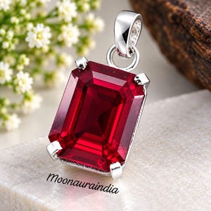 May include: A rectangular emerald-cut deep red gemstone pendant set in a polished silver-toned metal prong setting with a circular bail at the top. The text Moonauraindia is printed in black script on the surface below the pendant.