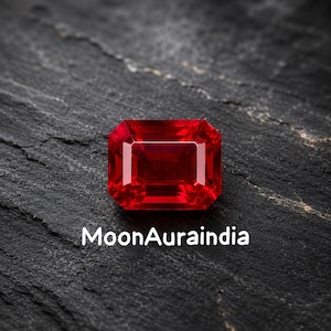 May include: A vibrant, rectangular-cut ruby gemstone with a deep red hue. The gem is set against a dark, textured surface. The text "MoonAuraindia" is displayed in white font below the gemstone.