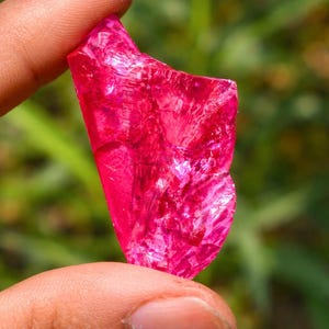 May include: A vivid, raw, pink gemstone held between fingers. The irregular shape of the stone catches the light, displaying a crystalline structure. The background is a soft, blurred green.