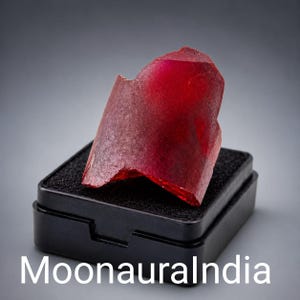 May include: A raw, deep red crystal specimen with a translucent, jagged structure. The stone is positioned upright on a black foam insert inside a small, square black plastic display box. The text MoonauraIndia is visible at the bottom.