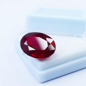 May include: A faceted, oval-shaped ruby gemstone. The gem is a deep red color with bright, reflective facets. It sits in a light blue plastic jewelry box, against a white background. The gemstone is a focal point, showcasing its clarity and brilliance.