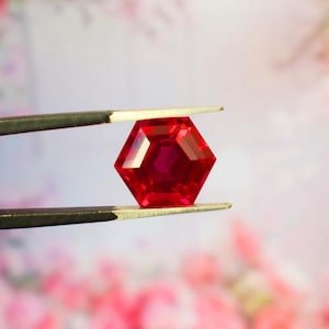 Puede incluir: Una gema facetada, hexagonal, de color rojo rubí es sostenida por unas pinzas. La gema tiene un tono rojo intenso y un diseño complejo y multifacético. El fondo es una suave mezcla borrosa de rosa y blanco.