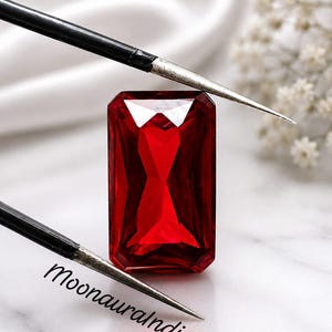 Pode incluir: Uma pedra preciosa retangular, facetada e de cor vermelho profundo, posicionada entre duas ferramentas metálicas finas com cabos pretos. A pedra exibe cortes geométricos nítidos que refletem a luz, com o texto MoonauraIndia impresso em preto.