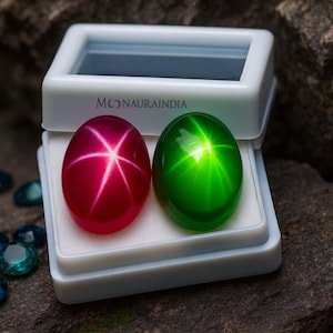 Puede incluir: Dos gemas ovaladas, una de color rojo rubí y otra verde esmeralda, cada una con un efecto de estrella. Se presentan en una vitrina blanca con el texto "MONAURAINDIA". Hay gemas facetadas adicionales dispersas cerca.