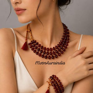 Könnte beinhalten: Ein mehrreihiges Halsketten- und Armband-Set. Der Schmuck besteht aus dunkelroten, tropfenförmigen Perlen. Die Halskette und das Armband haben goldfarbene Akzente und rote Quasten. Der Text "MoonAuraindia" ist im Bild zu sehen.