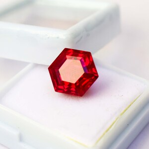 May include: A faceted, hexagonal red gemstone sits in a white display box. The gem has a central, lighter-coloured hexagon within the larger shape. The box is open, with the lid partially visible in the background.