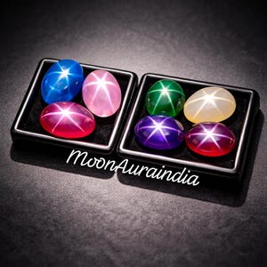Op de afbeelding: Twee zwarte displaydoosjes met acht ovale cabochon edelstenen, elk met een duidelijk zesstralig ster-effect. De stenen zijn blauw, roze, rood, groen, crème, paars en donkerrood. De tekst MoonAuraindia is in het wit weergegeven.