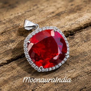 May include: A silver pendant featuring a large, square-cut, vibrant red gemstone at the center, surrounded by a halo of small, clear, sparkling stones. The pendant includes a silver bail at the top and displays the text MoonauraIndia below it.