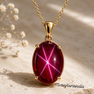 May include: A gold pendant necklace featuring an oval-shaped deep red gemstone with a prominent six-rayed star effect. The stone is held in a four-prong gold setting attached to a delicate gold chain. The text MoonAuraindia is visible.