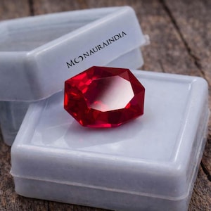 Może przedstawiać: Fasetowany, głęboko czerwony kamień szlachetny spoczywa na białym, plastikowym pudełku ekspozycyjnym. Pudełko jest otwarte, ukazując blask klejnotu. Na pudełku widnieje napis "MONAURAINDIA". Tło to rustykalna drewniana powierzchnia.