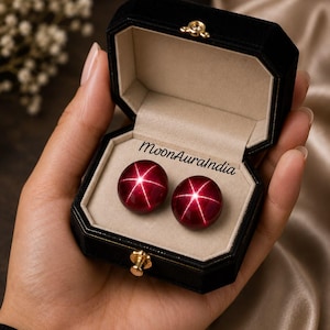 Pode incluir: Duas pedras preciosas cabochão redondas de cor vermelho profundo com um distinto efeito de estrela de seis pontas, apresentadas dentro de uma caixa de joias octogonal forrada com veludo preto. O interior da tampa exibe o texto MoonAuraIndia.