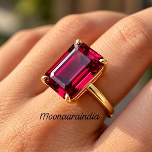 Könnte beinhalten: Ein Goldring mit einem großen, rechteckigen, smaragdgeschliffenen tiefroten Edelstein, der von vier Krappen gehalten wird. Die Ringschiene ist dünn und aus poliertem Gold. Der Text Moonauraindia ist in schwarzer Schrift unter dem Stein sichtbar.