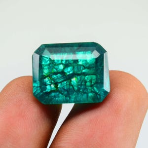 May include: Emerald-cut gemstone with a vibrant teal color and internal crystalline patterns. The gem is held between fingers against a white background. The facets of the stone reflect light, creating a sparkling effect.