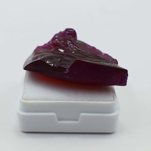May include: A deep magenta, translucent gemstone with an irregular, angular shape and faceted edges. The stone displays a rich, saturated purple-red hue with visible internal clarity and light refraction across its polished, uneven surface.