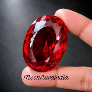 May include: A large, faceted, oval-shaped red gemstone. The gem is held between two fingers against a dark background. The surface of the gem reflects light, creating bright highlights. The text "MoonAuraindia" is visible on the finger.