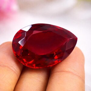Può includere: Una grande gemma sfaccettata a forma di pera, di colore rosso intenso. La superficie della gemma riflette la luce, mostrando le sue numerose sfaccettature. La pietra è tenuta in mano, evidenziando le sue dimensioni e il suo colore. Questa gemma è adatta alla creazione di gioielli.
