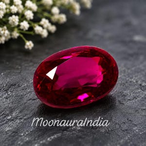 May include: A vibrant, oval-cut ruby gemstone with deep red coloring and multifaceted surfaces that reflect light. The gemstone is positioned on a dark, textured surface with the text MoonauraIndia printed in white script below the stone.
