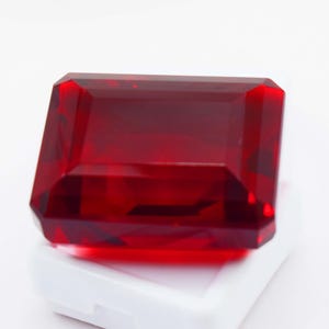 Puede incluir: Una piedra preciosa rectangular de color rojo intenso con talla esmeralda, que presenta múltiples facetas que reflejan la luz. La piedra es transparente y muestra un tono carmesí rico y saturado en toda su estructura, sobre una base blanca.