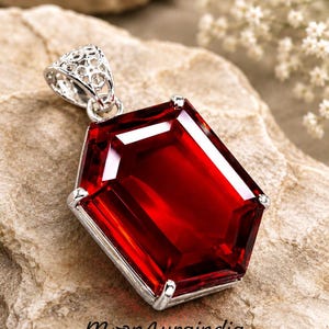 May include: A pendant featuring a large, faceted, deep red gemstone in a hexagonal shape, set in a silver-toned metal frame with a decorative, filigree-style bail. The text MoonAuraindia is printed in black at the bottom of the image.