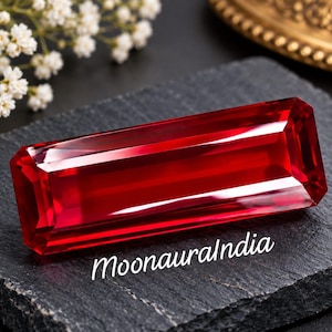 May include: A rectangular, faceted, deep red gemstone with sharp edges and a high-gloss finish. The stone is positioned on a dark, textured slate surface. The text "MoonauraIndia" is printed in white script on the slate below the gemstone.