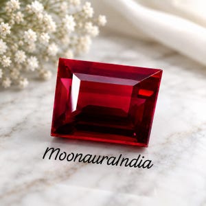 May include: A rectangular faceted gemstone with a deep, vibrant red colour and sharp, clean edges. The stone displays high clarity and internal light reflection. The text MoonauraIndia is printed in black script below the gemstone.