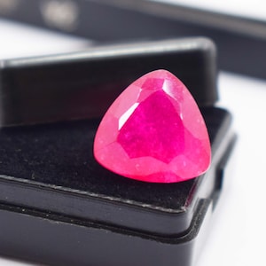 Könnte beinhalten: Ein leuchtend pinkfarbener Edelstein in dreieckigem Schliff mit zahlreichen Facetten, die das Licht reflektieren. Der Edelstein liegt in einer kleinen, schwarzen, rechteckigen Aufbewahrungsbox mit einem mit Samt ausgekleideten Innenraum.