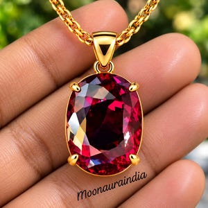 Puede incluir: Un collar con colgante de oro que presenta una gran piedra preciosa de color rojo intenso, de corte ovalado y sujeta por cuatro garras. El colgante cuelga de una cadena de oro gruesa. El texto "Moonauraindia" aparece en letras negras.