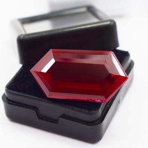 Puede incluir: Una piedra preciosa de color rojo intenso con facetas y forma hexagonal que descansa sobre una caja de presentación forrada de terciopelo negro. La piedra presenta bordes geométricos afilados y una superficie pulida que refleja la luz, contrastando con el contenedor rectangular oscuro.