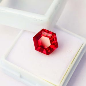 May include: A faceted, hexagonal red gemstone, displayed in a white case. The gem has a bright, reflective surface and a complex internal structure. The case is open, revealing the gem's vibrant color and intricate cut.
