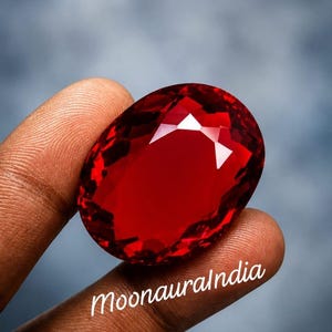 Könnte beinhalten: Ein großer, oval geschliffener, tiefroter Edelstein mit einer facettierten Oberfläche, die das Licht reflektiert. Der Stein hat einen brillanten, transparenten karmesinroten Farbton und ist mit dem weißen Text MoonauraIndia im unteren Bereich versehen.