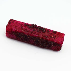 Könnte beinhalten: Ein rechteckiges Stück leuchtend magentafarbenes Drachenfruchtfleisch, das mit einer Schicht aus dunklen, groben, körnigen Gewürzen bedeckt ist. Das Fruchtsegment hat eine feste, blockartige Form mit einer texturierten Oberfläche.
