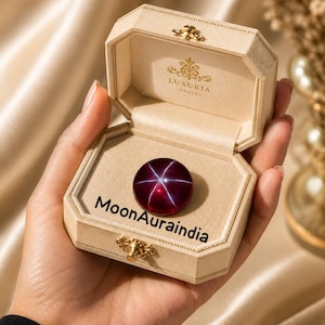 Op de afbeelding: Een ronde, dieprode edelsteen met een stervormig patroon wordt getoond in een open, crèmekleurige sieradendoos. De doos is voorzien van gouden accenten en de tekst "LUXURIA JEWELRY". De tekst "MoonAuraindia" is op de doos gedrukt.