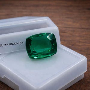 Op de afbeelding: Een levendige, smaragdgroene edelsteen ligt op een witte plastic doos. De edelsteen is kussenvormig geslepen en reflecteert licht. De doos is open en onthult een compartiment. De tekst "MONAURAINDIA" staat op de doos.