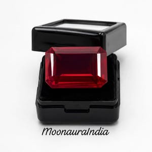Könnte beinhalten: Ein tiefroter Edelstein im Smaragdschliff, der in einer kleinen, schwarzen aufklappbaren Schmuckschatulle mit schwarzem Samtinnenfutter liegt. Der Text MoonauraIndia ist in Schwarz unter der Schatulle aufgedruckt. Der Edelstein hat klare Facetten.