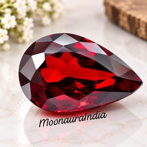 May include: A pear-shaped, faceted deep red gemstone with a brilliant cut that reflects light across its surface. The stone is positioned on a light surface with the text MoonauraIndia printed in black lettering below the gemstone.