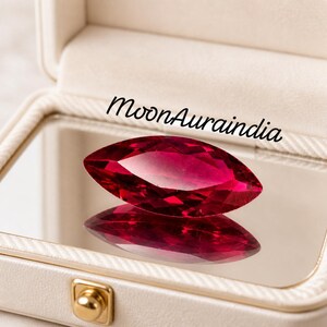 May include: A marquise-cut, ruby-red gemstone is displayed inside a white jewelry box with a mirrored base. The gemstone's facets reflect light, creating a brilliant sparkle. The text "MoonAuraindia" is written in black script.