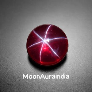 May include: A spherical, polished, deep red gemstone displaying a six-rayed star pattern. The gem is set against a dark grey background. The text "MoonAuraindia" is printed in white at the bottom of the image.