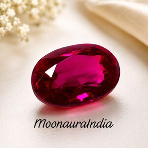Op de afbeelding: Een ovaal geslepen, diep magentarode edelsteen met meerdere facetten die het licht reflecteren. De steen bevindt zich boven de zwarte tekst MoonauraIndia, die in een cursief lettertype op een glad, lichtgekleurd oppervlak is gedrukt.
