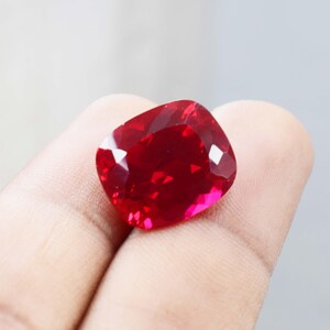 Könnte beinhalten: Ein facettierter Rubin-Edelstein im Kissenschliff. Der Edelstein ist ein tiefes, leuchtendes Rot mit hellen Glanzlichtern. Die Form ist ein modifiziertes Quadrat mit abgerundeten Ecken, das die Brillanz und Klarheit des Steins zeigt. Der Rubin wird in einer Hand gehalten.