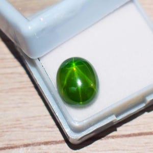 Pode incluir: Uma vibrante pedra preciosa verde com um padrão em forma de estrela, apresentada sobre um fundo branco dentro de uma caixa de plástico azul claro. A superfície polida reflete a luz, mostrando a clareza e a cor da gema. Esta pedra preciosa é um belo exemplo de joalharia natural.